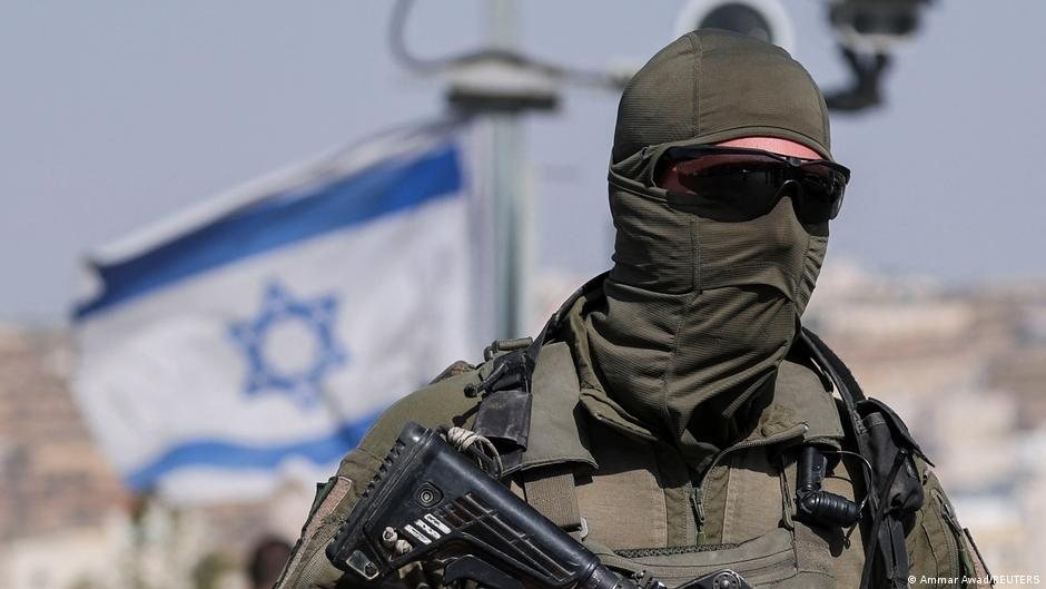 Un soldado con uniforme táctico, pasamontañas y gafas de sol sostiene un fusil frente a una bandera de Israel