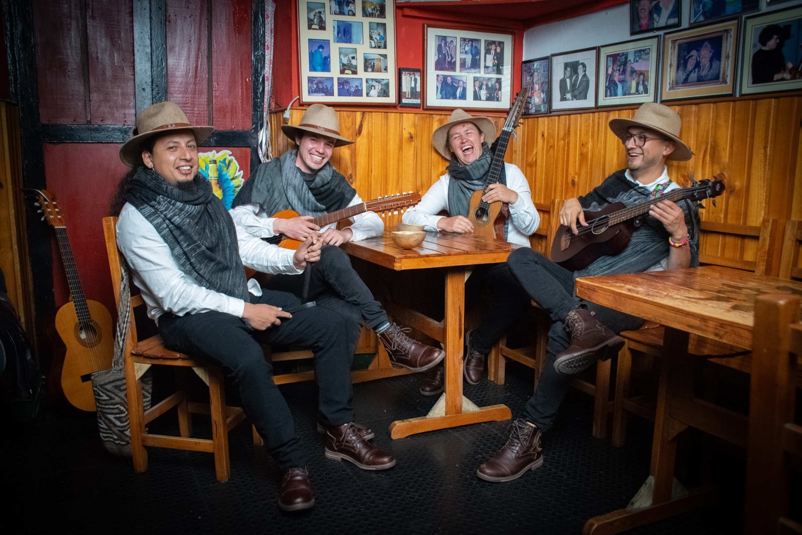 Cuatro músicos con sombreros y guitarras sentados en mesas de madera dentro de un local rústico
