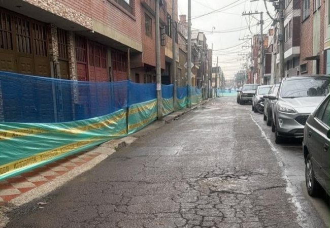 Calle con pavimento deteriorado y una malla de construcción azul y amarilla frente a edificios de ladrillo
