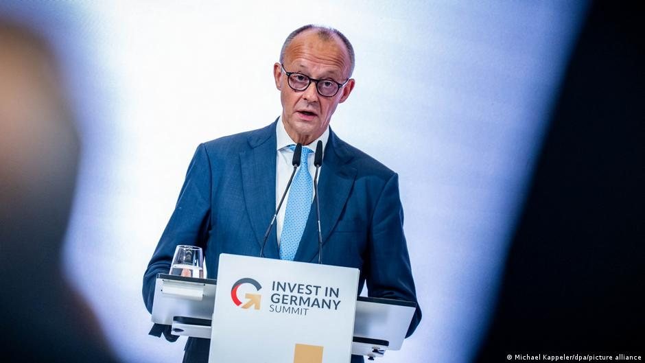 Un hombre con traje azul y gafas habla frente a un podio con el logo de Invest in Germany Summit