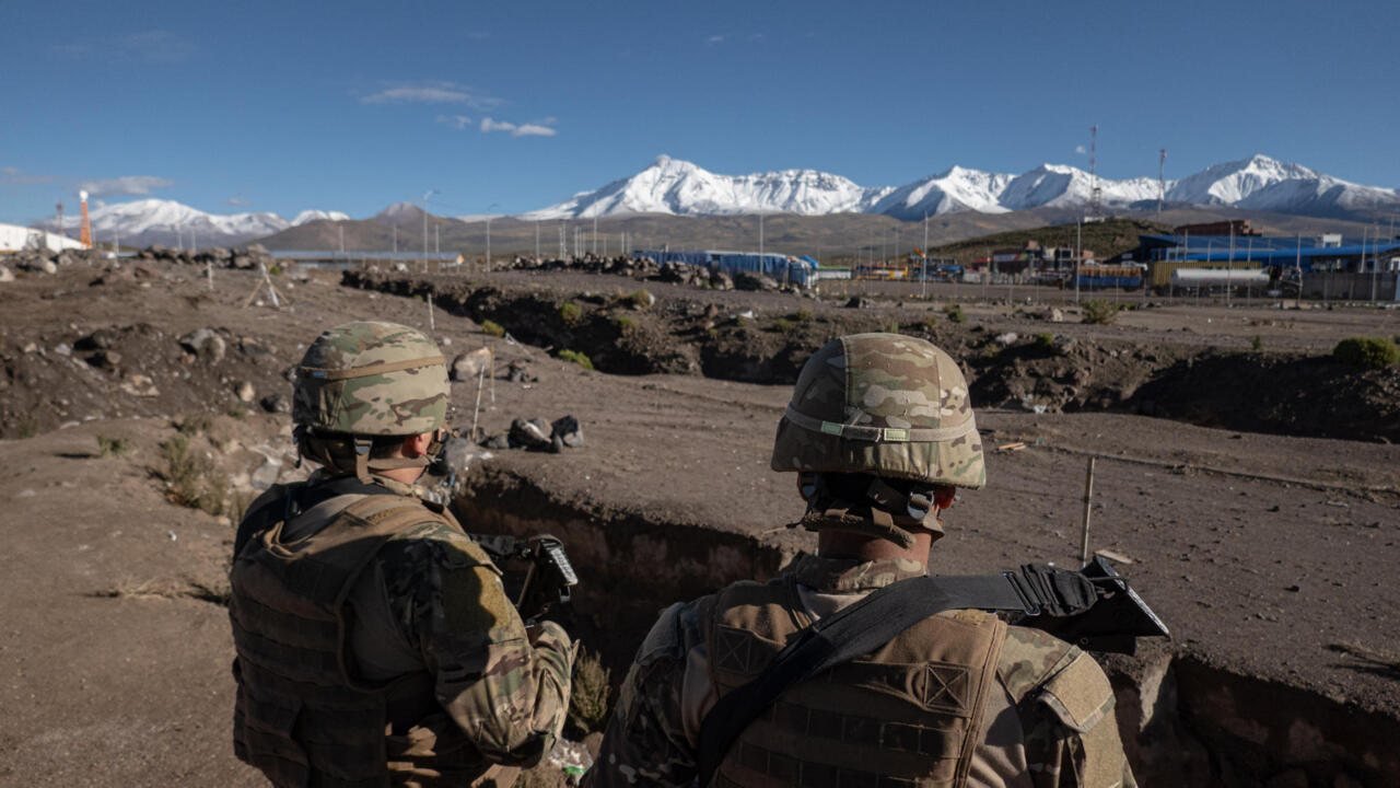 Dos soldados con equipo táctico y cascos camuflados observan un paisaje desértico con montañas nevadas al fondo