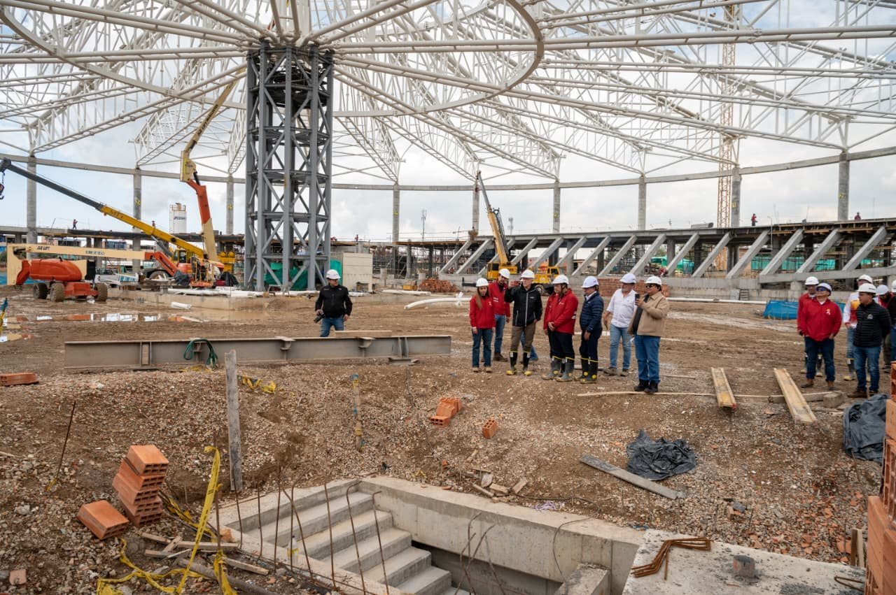 Un grupo de personas con cascos de seguridad y chalecos frente a la estructura metálica de un velódromo en construcción