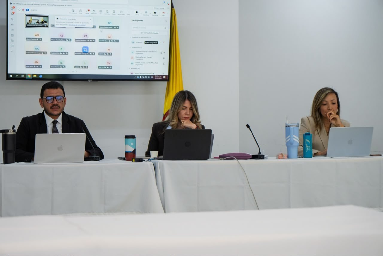 Tres personas sentadas frente a computadores en una mesa larga durante una audiencia pública