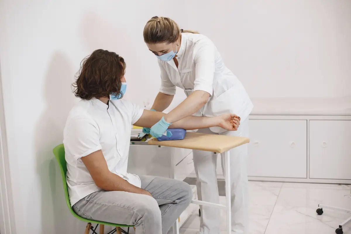 Una enfermera con mascarilla y guantes azules toma la presión arterial a un hombre sentado en una silla