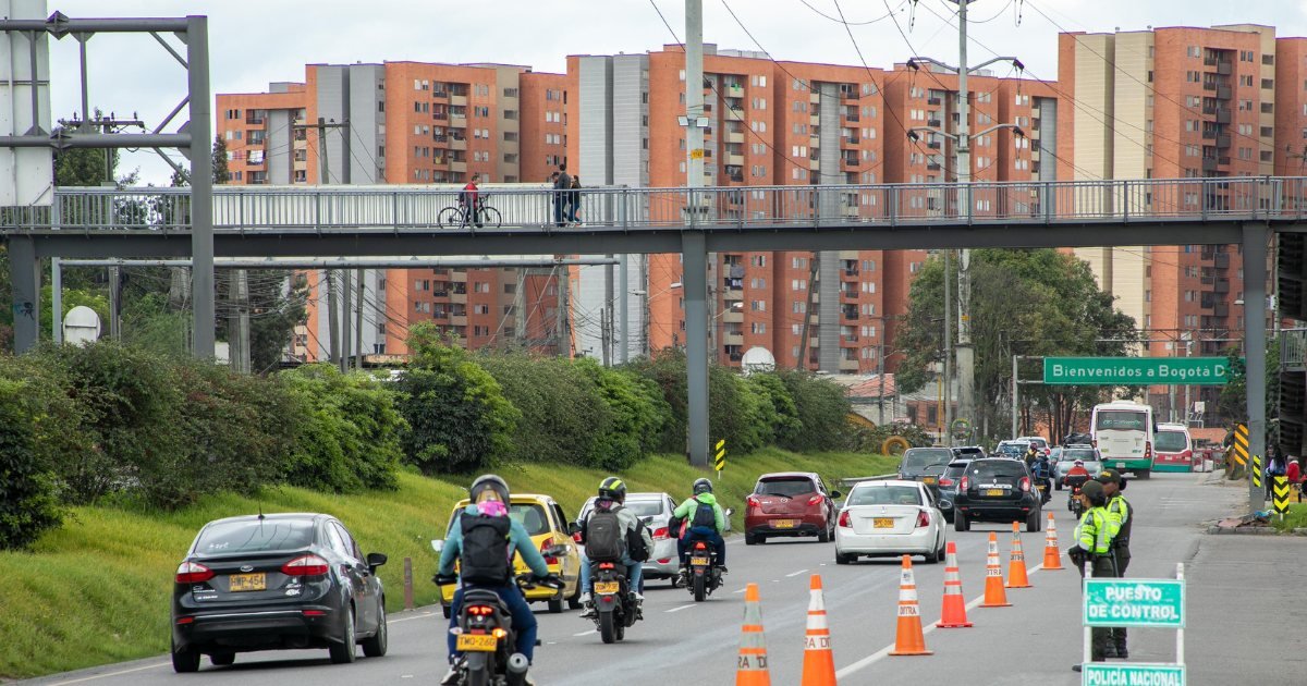 Vehículos y motocicletas circulan por una vía en Bogotá con un puente peatonal y edificios de apartamentos al fondo