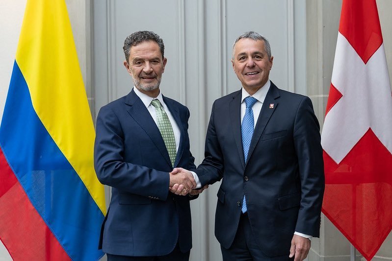 Dos hombres con traje de negocios se dan la mano frente a las banderas de Colombia y Suiza