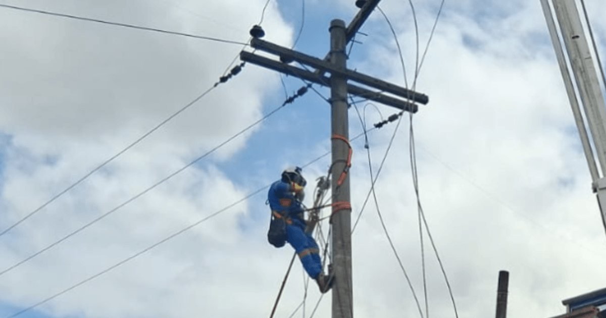 Un trabajador de mantenimiento eléctrico con equipo de seguridad subido a un poste de alta tensión bajo un cielo nublado.