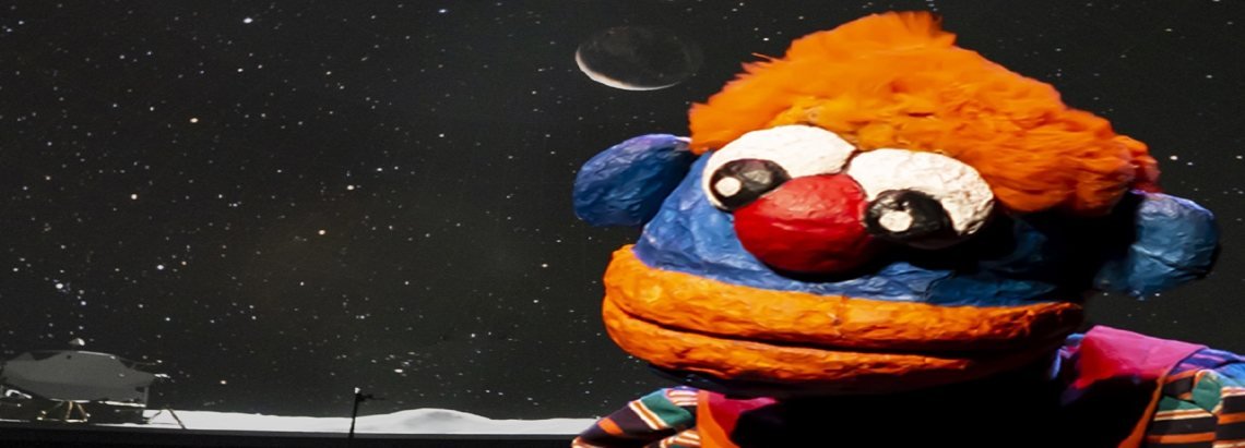 Un títere de color azul y naranja con ojos grandes frente a un fondo de estrellas y la luna