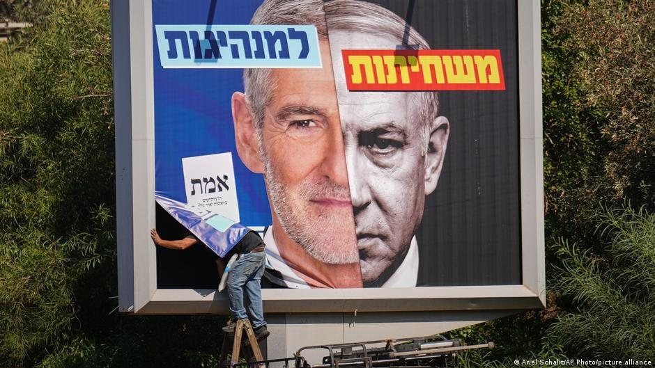 Una persona en una escalera coloca un cartel sobre una valla publicitaria con rostros de políticos en Israel