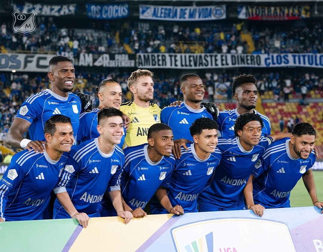 Jugadores de fútbol con uniformes azules posan juntos en un estadio frente a una barrera publicitaria