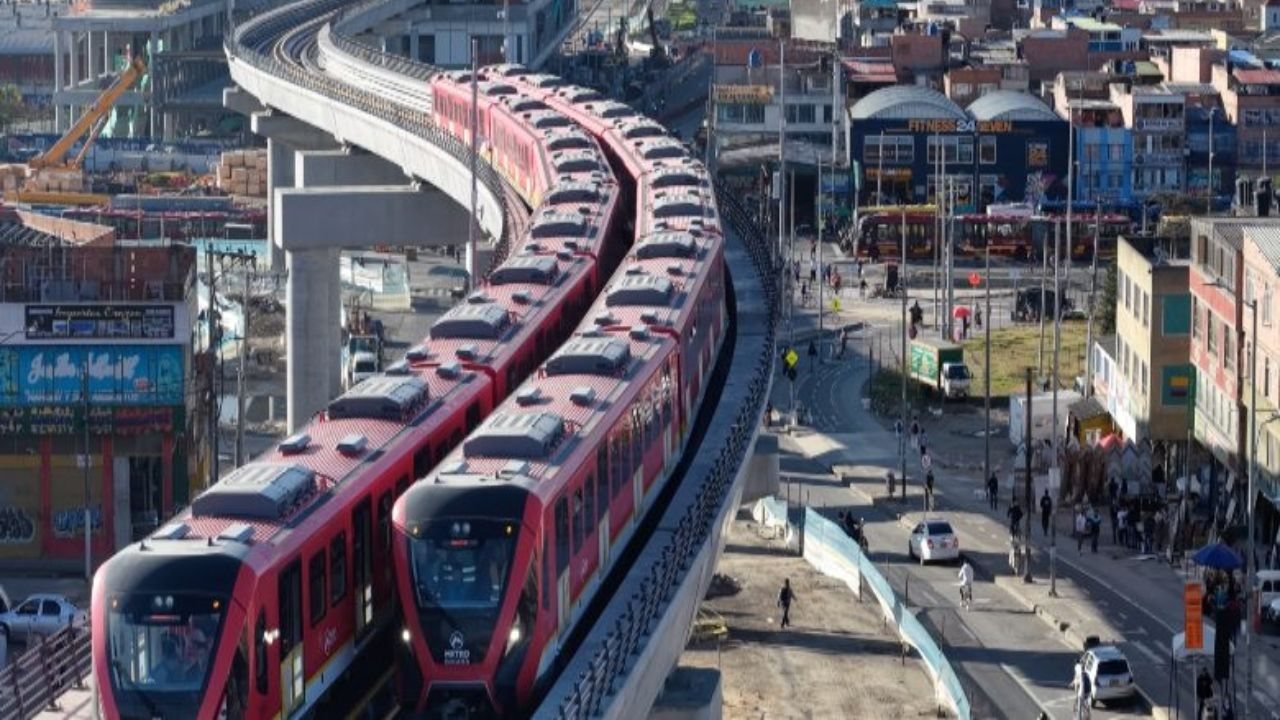 Dos trenes rojos circulan por una vía elevada en una zona urbana con edificios y comercio