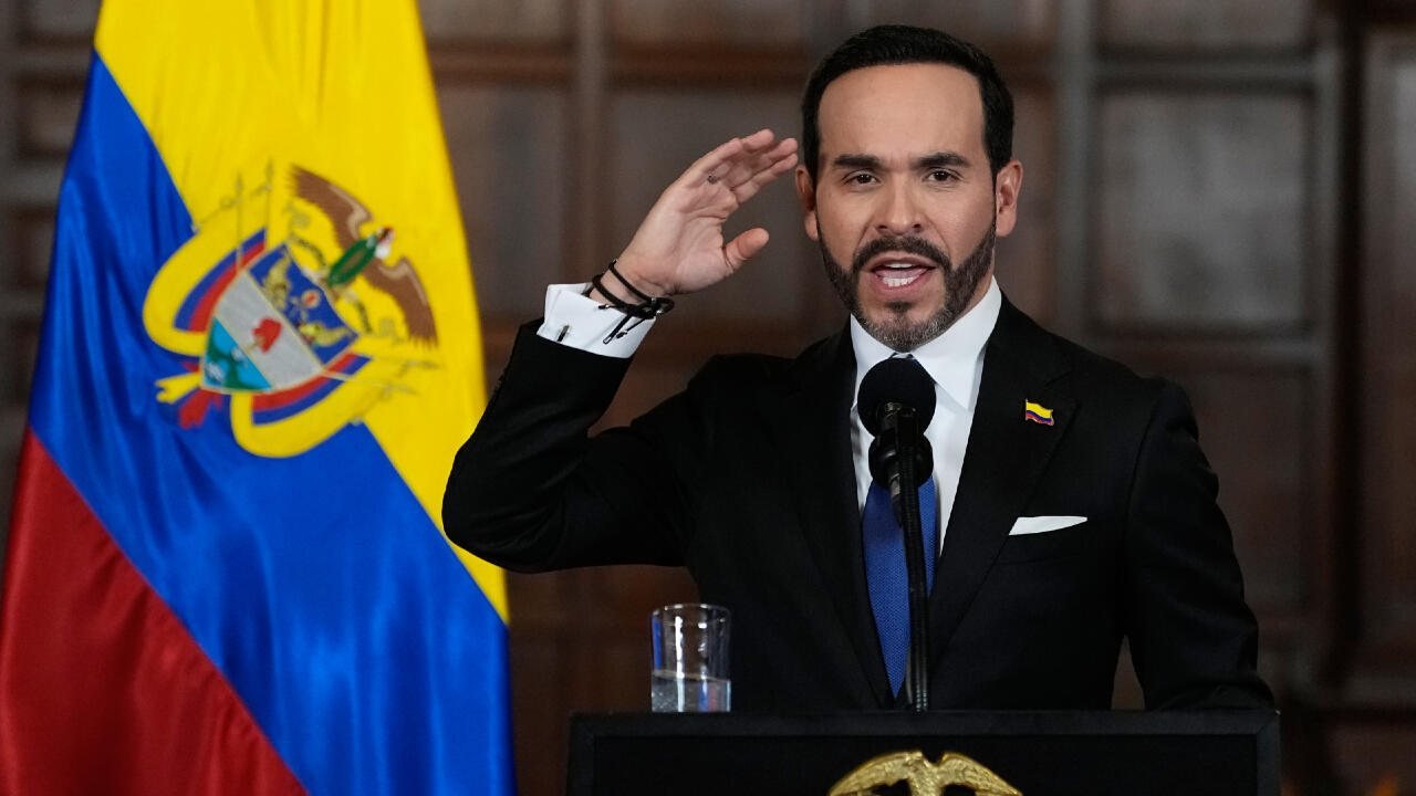Un hombre con traje oscuro y corbata azul habla frente a un micrófono con la bandera de Colombia al fondo