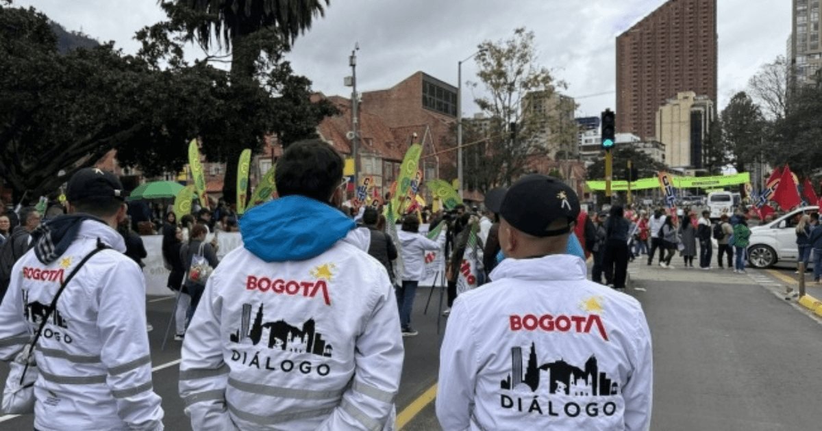 Tres personas de espaldas visten chaquetas blancas con el logo de Bogotá y la palabra Diálogo en una calle con gente