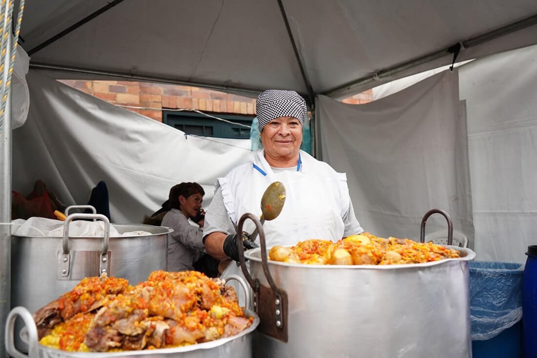 Un hombre con gorro de cocina y delantal sostiene una cuchara grande frente a ollas de metal con comida en una carpa