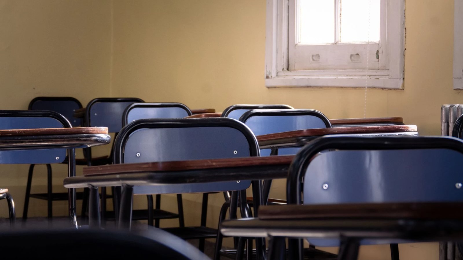 Sillas y pupitres azules en un salón de clases vacío con paredes amarillas y una ventana