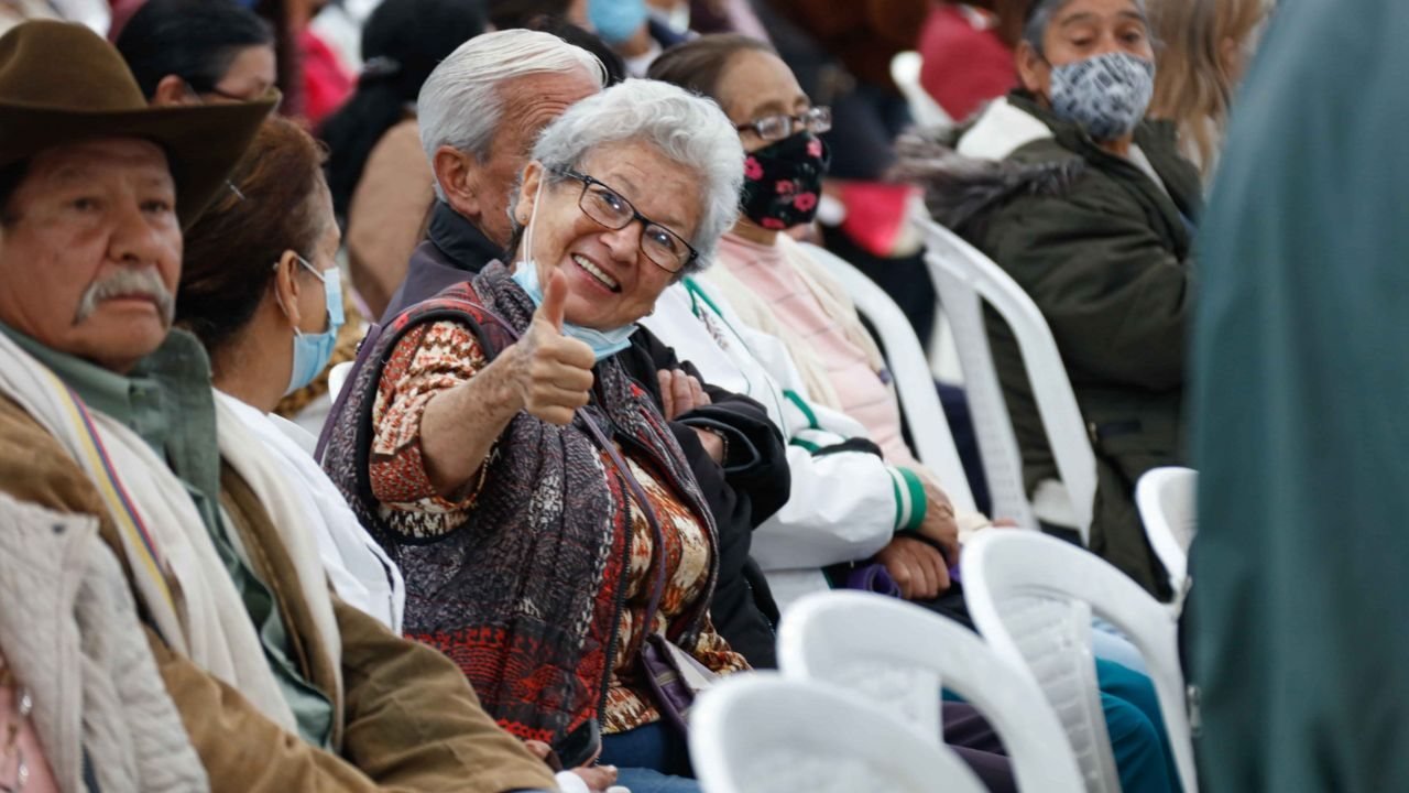 Una mujer mayor con gafas y una manta tradicional sonríe y levanta el pulgar hacia la cámara en un evento