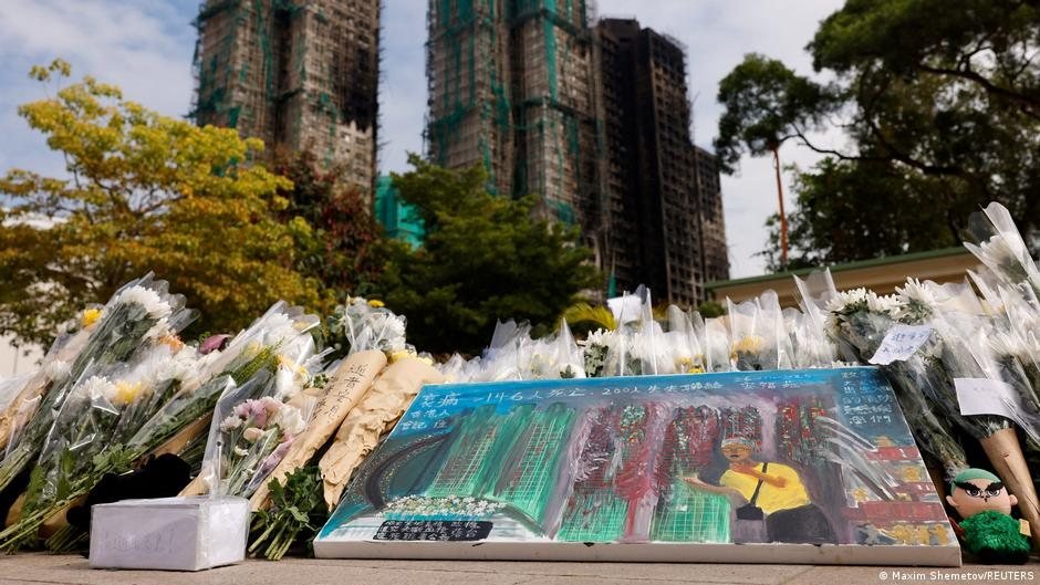 Ramos de flores blancas y un cartel conmemorativo en una calle frente a edificios en construcción