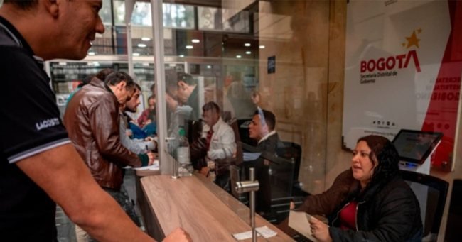 Personas en una oficina de atención al ciudadano con un mostrador de madera y un cartel de Bogotá