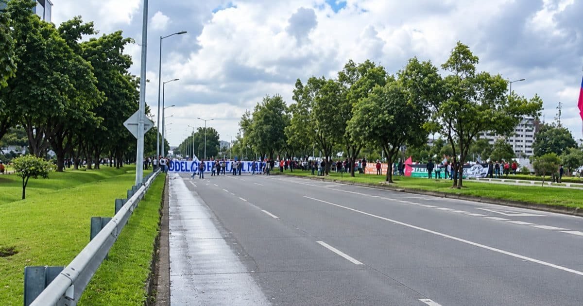 Manifestantes marchan por una avenida principal bajo un cielo parcialmente nublado.