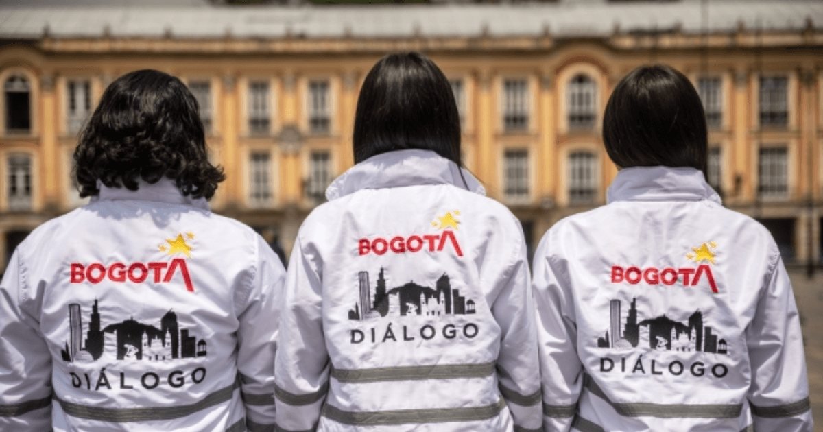 Tres personas de espaldas visten uniformes blancos con el logo de 'Bogotá Diálogo' frente a un edificio histórico.