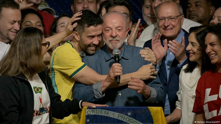 Un hombre con barba y camisa azul es abrazado por un hombre con camiseta de fútbol amarilla en un escenario