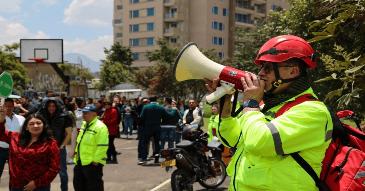 Un hombre con casco y chaleco reflectante usa un megáfono en una plaza pública rodeado de personas.