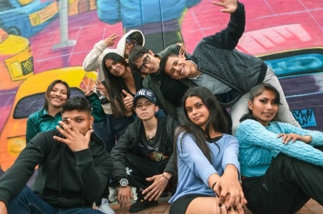 Un grupo de jóvenes posa sonriendo frente a un mural colorido en una calle de Bogotá