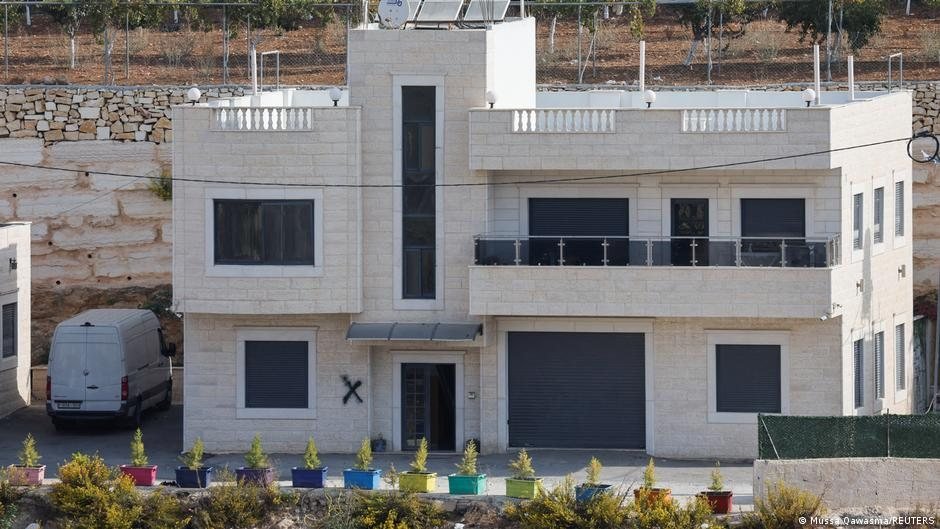 Fachada de una casa de piedra blanca con ventanas oscuras y una furgoneta gris estacionada frente a ella
