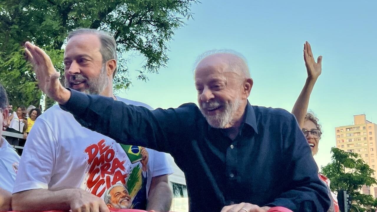 Dos hombres sonríen y saludan a la cámara en un evento al aire libre con árboles y edificios de fondo