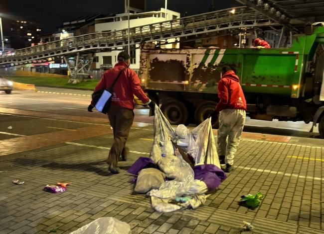 Dos personas con chaquetas rojas recogen bolsas de basura junto a un camión recolector en una calle de noche