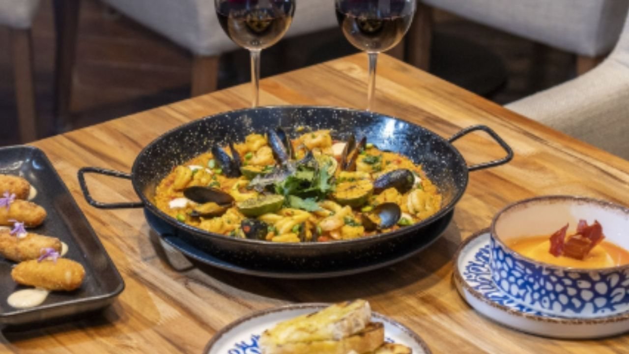 Una mesa de madera con una paella de mariscos, copas de vino y otros platos de comida en un restaurante