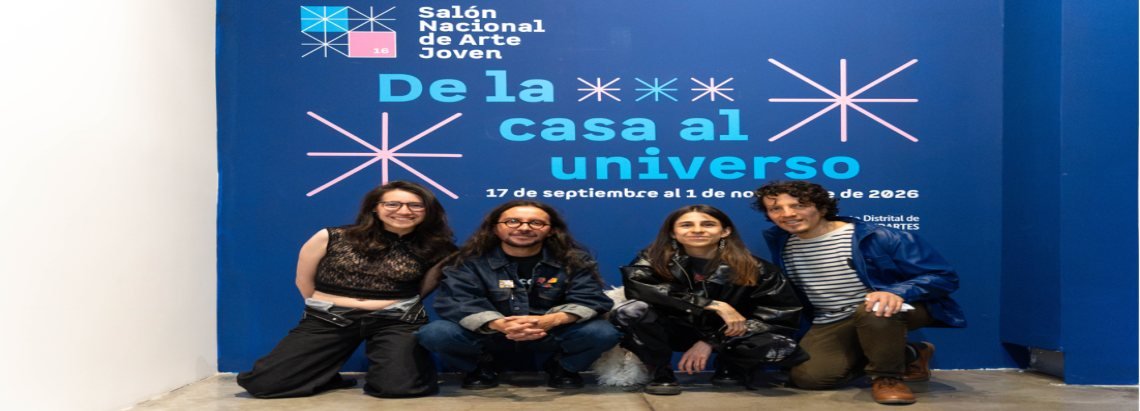 Cuatro personas posan sentadas frente a un muro azul con el texto de la exposición De la casa al universo