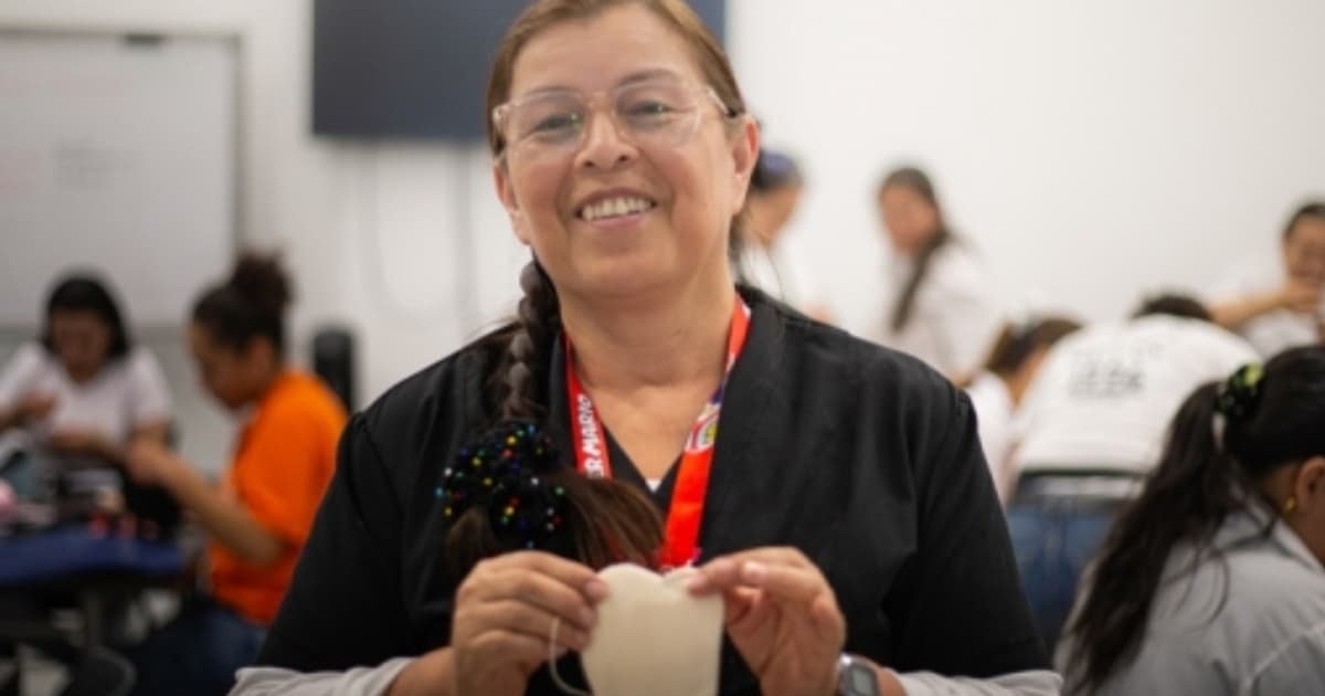 Una mujer sonriente con gafas y una acreditación roja sostiene un objeto pequeño en un entorno de trabajo.